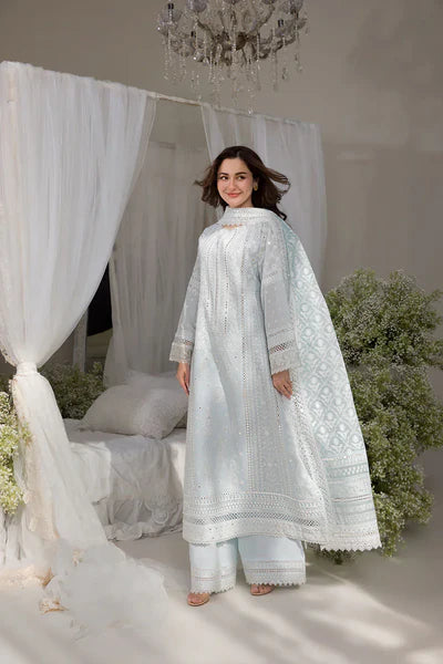 Woman in a white embroidered traditional outfit standing in a decorated room with white curtains and plants.