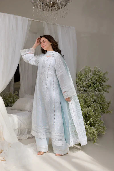 Woman in a white embroidered outfit standing in a softly lit room with white curtains and greenery.